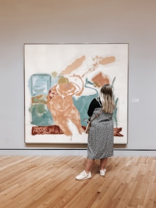 A person with long hair stands in front of an abstract painting in a gallery. The painting features large, expressive brushstrokes in shades of blue, orange, and white. The gallery room has wooden floors and neutral-colored walls, enhancing the focus on the artwork.