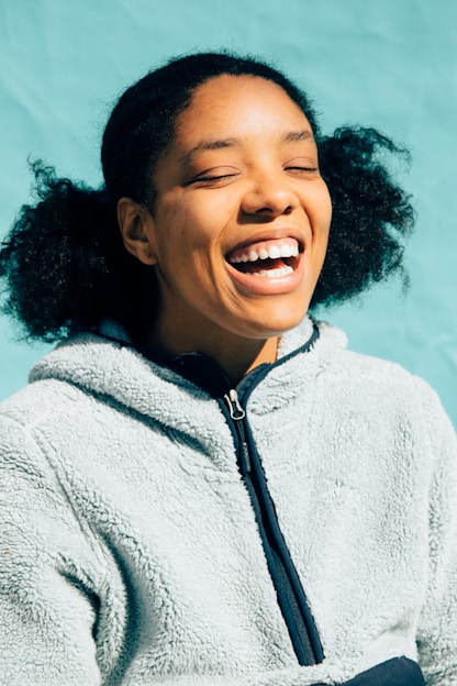 A person with a joyful expression, eyes closed, and mouth open in laughter. They are wearing a fuzzy light-colored jacket against a blue background.