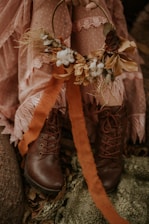 A person is holding a decorative autumn-themed wreath made of dried flowers, cotton, and leaves. They are dressed in a lacy, pink outfit with polka dots and wearing brown leather boots. The setting has an earthy and rustic feel, with fallen leaves and a green textured fabric on the ground.