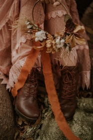 A person is holding a decorative autumn-themed wreath made of dried flowers, cotton, and leaves. They are dressed in a lacy, pink outfit with polka dots and wearing brown leather boots. The setting has an earthy and rustic feel, with fallen leaves and a green textured fabric on the ground.
