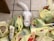 A white cat is surrounded by several yellow plastic grocery bags scattered on a tiled floor. Various food items are partially visible within the bags, including boxes and jars. The cat looks up at the camera with a curious expression.
