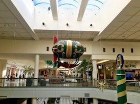 A shopping mall interior with a large decorative blimp suspended from the ceiling, featuring ornate designs and red accents. The mall includes various stores with visible signage, including clothing retailers. Several potted plants and decorative star-shaped ornaments are also present.
