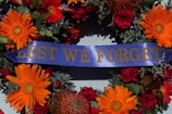 A commemorative wreath featuring vibrant orange and red flowers, with accents of green foliage and a central blue ribbon displaying the words 'LEST WE FORGET' in gold lettering.