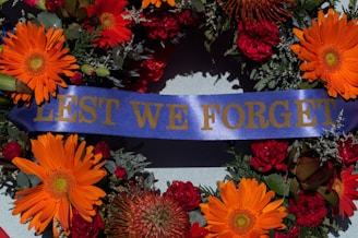 A commemorative wreath featuring vibrant orange and red flowers, with accents of green foliage and a central blue ribbon displaying the words 'LEST WE FORGET' in gold lettering.