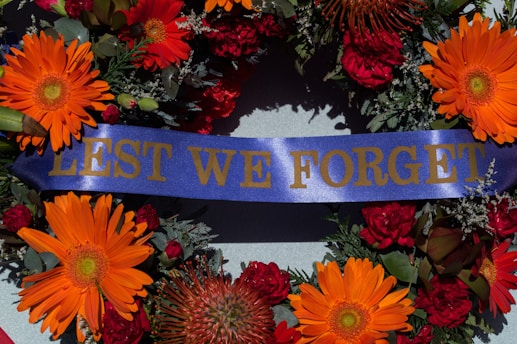 A commemorative wreath featuring vibrant orange and red flowers, with accents of green foliage and a central blue ribbon displaying the words 'LEST WE FORGET' in gold lettering.