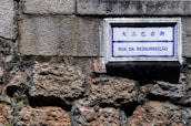 A street sign with the words 'Rua da Ressurreição' is mounted on a textured stone wall. The sign has both Chinese and Portuguese text, with a blue and white color scheme.