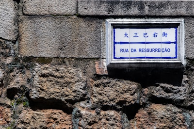 A street sign with the words 'Rua da Ressurreição' is mounted on a textured stone wall. The sign has both Chinese and Portuguese text, with a blue and white color scheme.