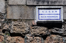 A street sign with the words 'Rua da Ressurreição' is mounted on a textured stone wall. The sign has both Chinese and Portuguese text, with a blue and white color scheme.