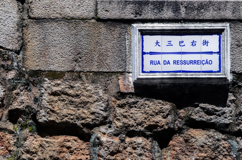 A street sign with the words 'Rua da Ressurreição' is mounted on a textured stone wall. The sign has both Chinese and Portuguese text, with a blue and white color scheme.