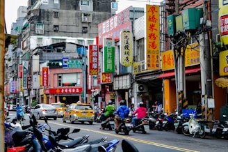 A busy urban street with multiple motorcycles lined up along the sidewalk and several cars, including taxis, on the road. Buildings are adorned with numerous large, colorful signs in Chinese characters. The scene includes pedestrians, some on motorcycles, amidst an atmosphere typical of a bustling city.