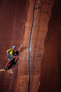 A climber wearing a helmet and harness stands on a narrow ledge against a tall red sandstone wall. The climber is geared up with climbing equipment and ropes, which are secured above. The rock face features a prominent crack running vertically, emphasizing the challenging nature of the climb.