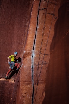 A climber wearing a helmet and harness stands on a narrow ledge against a tall red sandstone wall. The climber is geared up with climbing equipment and ropes, which are secured above. The rock face features a prominent crack running vertically, emphasizing the challenging nature of the climb.