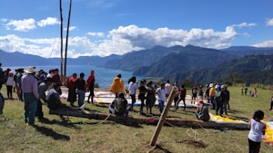 A scenic outdoor gathering of people on a grassy hilltop with a stunning view of mountains and a lake in the background. The group appears to be setting up or working on a project with colorful materials spread on the ground. The sky is mostly clear with fluffy clouds, providing a picturesque backdrop.