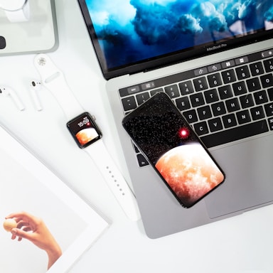A flat lay arrangement of technology and print media featuring a laptop with a cloudy sky wallpaper, a smartphone displaying a planet with a red spot, an Apple Watch with a similar screen, wireless earphones, and a magazine showing a hand holding a macaron.