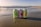 Three cans of Sanzo sparkling water are positioned on the wet sand of a beach with a calm ocean in the background. The scene includes distant mountains and city buildings under a clear blue sky, creating a serene coastal setting.