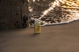 A can of Sanzo mango-flavored sparkling water sits on a sandy beach near the edge of the ocean. Gentle waves approach, capturing the golden light of a setting or rising sun, while a barnacle-covered pillar stands nearby.