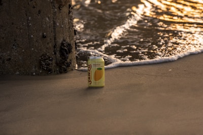 A can of Sanzo mango-flavored sparkling water sits on a sandy beach near the edge of the ocean. Gentle waves approach, capturing the golden light of a setting or rising sun, while a barnacle-covered pillar stands nearby.