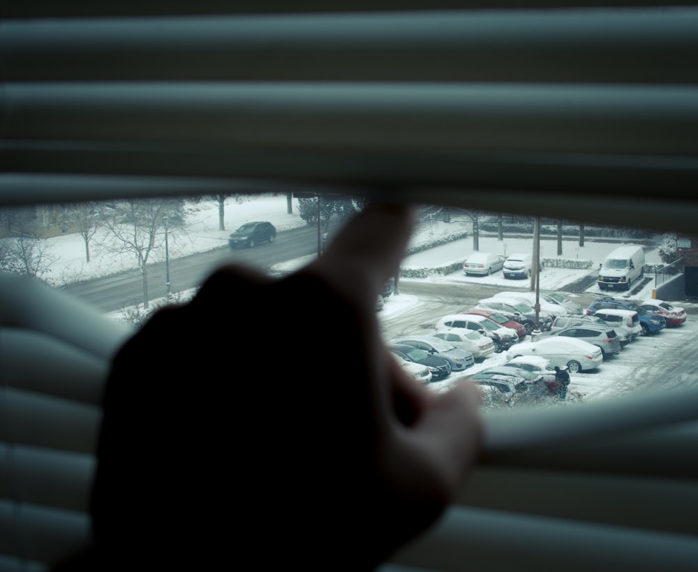 person using finger opening Venetian blinds with view of snow-covered cars parked on parking lot