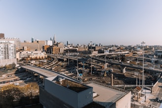 A sprawling urban landscape featuring a complex network of highways with multiple lanes, set against a backdrop of city skyscrapers. There are numerous vehicles traveling in both directions, and the scene captures the hustle and bustle of city life. Construction work is noticeable in some areas beside the highways, indicating ongoing development or repairs.