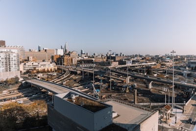 A sprawling urban landscape featuring a complex network of highways with multiple lanes, set against a backdrop of city skyscrapers. There are numerous vehicles traveling in both directions, and the scene captures the hustle and bustle of city life. Construction work is noticeable in some areas beside the highways, indicating ongoing development or repairs.
