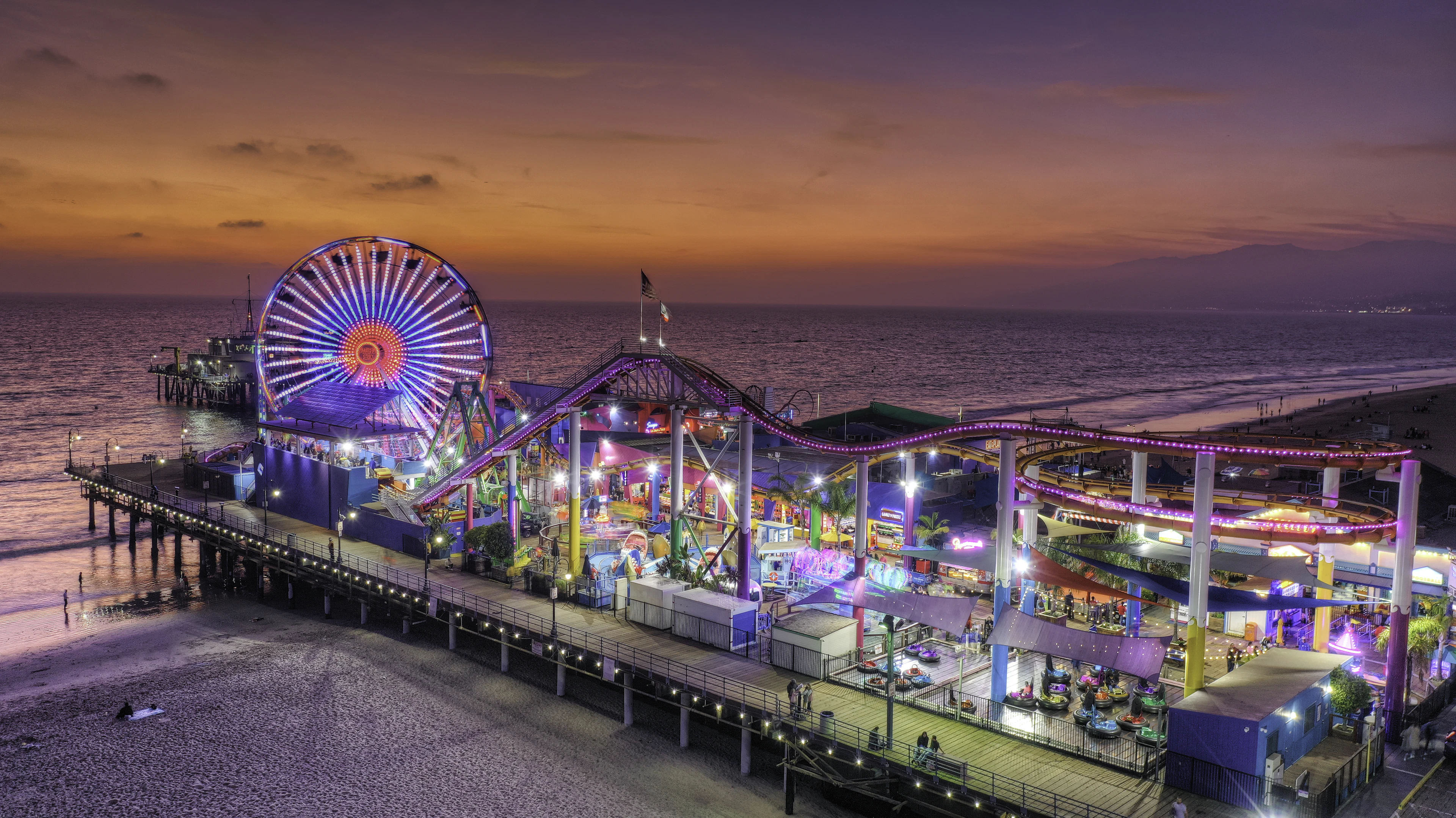 Santa Monica Pier at dusk, its Ferris wheel and roller coaster lit up in colour above the dark Pacific, under a violet and amber sky.