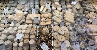 A diverse selection of cheeses displayed on a market counter, with various shapes, sizes, and textures. The cheeses are labeled with name tags and prices, and are arranged neatly. Some cheeses are wrapped in leaves, while others are round or cylindrical.