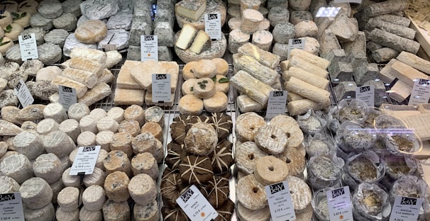 A diverse selection of cheeses displayed on a market counter, with various shapes, sizes, and textures. The cheeses are labeled with name tags and prices, and are arranged neatly. Some cheeses are wrapped in leaves, while others are round or cylindrical.