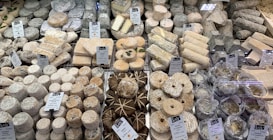A diverse selection of cheeses displayed on a market counter, with various shapes, sizes, and textures. The cheeses are labeled with name tags and prices, and are arranged neatly. Some cheeses are wrapped in leaves, while others are round or cylindrical.