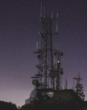 A silhouetted communication tower with numerous antennas and dishes stands against a starry night sky. The structure is tall and intricate, surrounded by a few smaller towers and some vegetation at the base.