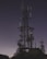 A silhouetted communication tower with numerous antennas and dishes stands against a starry night sky. The structure is tall and intricate, surrounded by a few smaller towers and some vegetation at the base.