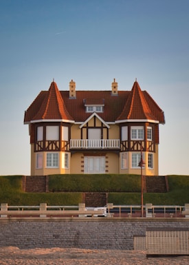 A picturesque house with a distinct architectural style, featuring red-tiled conical roofs and timber framing. It stands on a raised grassy mound with a stone retaining wall. The sky is clear, and a faint contrail is visible. A decorative lamp post and a stone fence are present in the foreground.