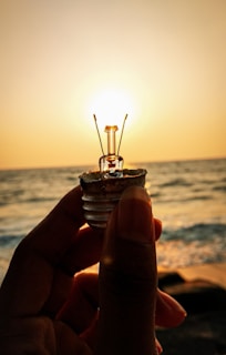 A person holds up a small light bulb against a backdrop of an ocean or sea at sunset. The sun is positioned directly behind the bulb, creating an effect where the bulb appears to be lit by the sunlight. The water below reflects the warm hues of the sunset.