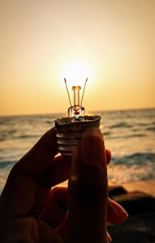 A person holds up a small light bulb against a backdrop of an ocean or sea at sunset. The sun is positioned directly behind the bulb, creating an effect where the bulb appears to be lit by the sunlight. The water below reflects the warm hues of the sunset.