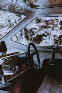 A well-designed boat cockpit area detailed with wooden paneling, featuring a steering wheel and various controls. The brand name 'Suzuki' is visible on one of the components. The seating area is upholstered in brown leather, creating a vintage aesthetic. Through the windows, out-of-focus trees can be seen, suggesting a natural outdoor setting.
