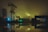 A nighttime scene with a foggy atmosphere, featuring silhouettes of trees and industrial buildings arranged around a large puddle reflecting light from nearby structures. A tower and several light sources cast a green and blue glow across the area, creating an eerie, mysterious ambiance.