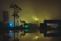 A nighttime scene with a foggy atmosphere, featuring silhouettes of trees and industrial buildings arranged around a large puddle reflecting light from nearby structures. A tower and several light sources cast a green and blue glow across the area, creating an eerie, mysterious ambiance.