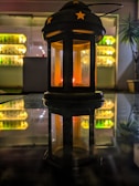A black lantern with star-shaped cutouts and a warm glowing light inside sits on a reflective surface. Behind it, shelves filled with bottles are softly illuminated, creating a cozy and inviting atmosphere. A plant with slender leaves is visible to the right.