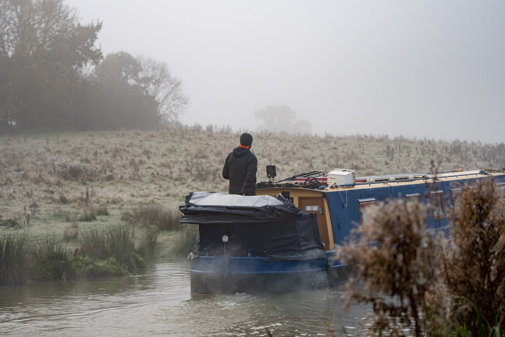 man wearing black and red jacket standing on blue boat near green field in foggy day