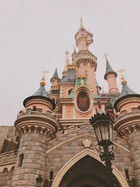A fairytale castle featuring ornate architecture with tall spires, vibrant stained glass, and intricate stonework. The design includes colorful turrets and golden accents, creating an enchanting and whimsical appearance. A black lantern stands in front, adding to the historic and magical atmosphere.