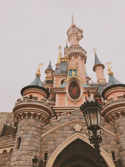 A fairytale castle featuring ornate architecture with tall spires, vibrant stained glass, and intricate stonework. The design includes colorful turrets and golden accents, creating an enchanting and whimsical appearance. A black lantern stands in front, adding to the historic and magical atmosphere.