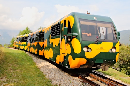 A train with a distinctive green and yellow camouflage pattern travels along a track surrounded by lush greenery and mountainous landscape. The train has a sleek, modern design and appears to be on a scenic route. A person is visible inside the driver's cabin.