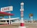 A vintage-style cafe and motel situated in a desert landscape with a clear blue sky. The cafe features a red and white color scheme with a visible sign displaying the word 'CAFE'. In front of the cafe, there is an old-fashioned gasoline price sign listing prices for unleaded, premium, and diesel fuel. To the right, a large sign for 'Roy's Motel Cafe' stands prominently. The background shows a row of white motel units and a mountainous desert horizon.