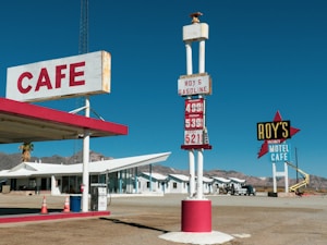 A vintage-style cafe and motel situated in a desert landscape with a clear blue sky. The cafe features a red and white color scheme with a visible sign displaying the word 'CAFE'. In front of the cafe, there is an old-fashioned gasoline price sign listing prices for unleaded, premium, and diesel fuel. To the right, a large sign for 'Roy's Motel Cafe' stands prominently. The background shows a row of white motel units and a mountainous desert horizon.
