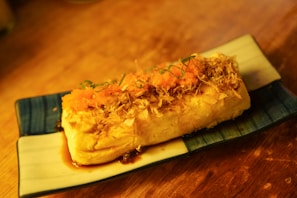 A rectangular piece of tofu topped with vibrant orange fish roe, garnished with thin slices of green onion and bonito flakes, served on a rectangular dish with a blue and white pattern. The dish sits on a wooden surface.