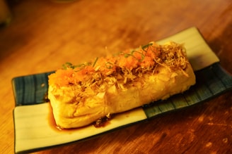 A rectangular piece of tofu topped with vibrant orange fish roe, garnished with thin slices of green onion and bonito flakes, served on a rectangular dish with a blue and white pattern. The dish sits on a wooden surface.
