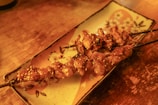 Grilled skewers of seasoned meat served on a decorative rectangular plate with floral patterns, resting on a wooden surface. The skewers appear to be sprinkled with sesame seeds.