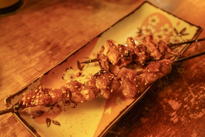 Grilled skewers of seasoned meat served on a decorative rectangular plate with floral patterns, resting on a wooden surface. The skewers appear to be sprinkled with sesame seeds.