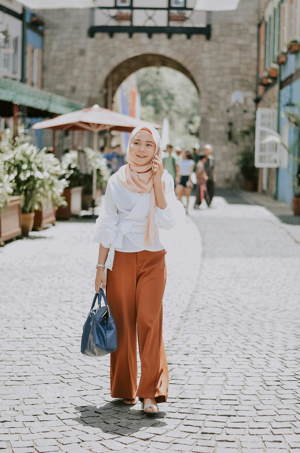 woman wearing beige hijab headscarf holding blue handbag using phone while walking on pathway