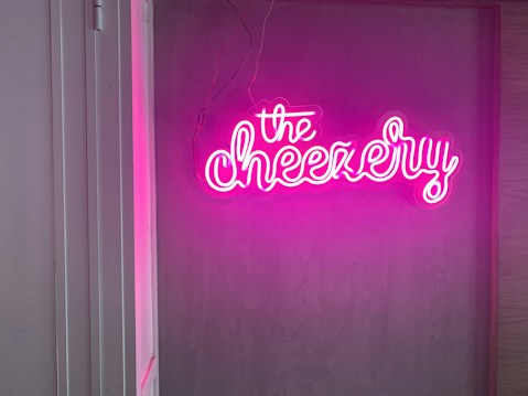 A glowing pink neon sign displaying the word 'the Cheezery' mounted on a plain wall, providing an eye-catching focal point. The sign's bright illumination contrasts with the muted tones of the wall.