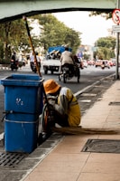 A person wearing a yellow uniform and orange hat is crouching beside a blue trash bin on a city sidewalk. A long broom is laying on the ground next to them. In the background, there is a busy street with vehicles and a bridge overhead. Trees line the side of the road amidst the urban setting.